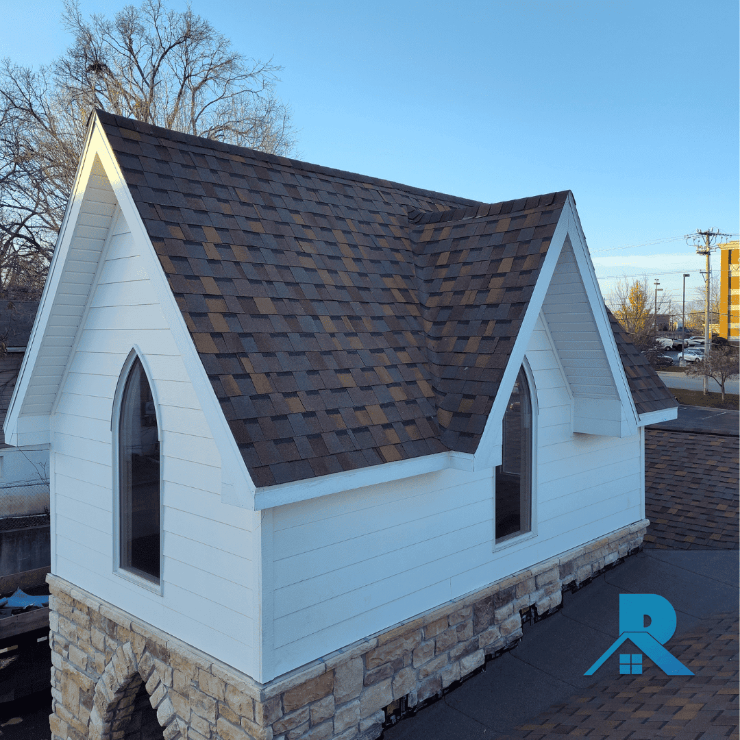 A small white chapel-style building with arched windows and a stone foundation, featuring a newly installed brown architectural shingle roof. The Branson Springfield Roof Co. logo appears in the bottom right corner.
